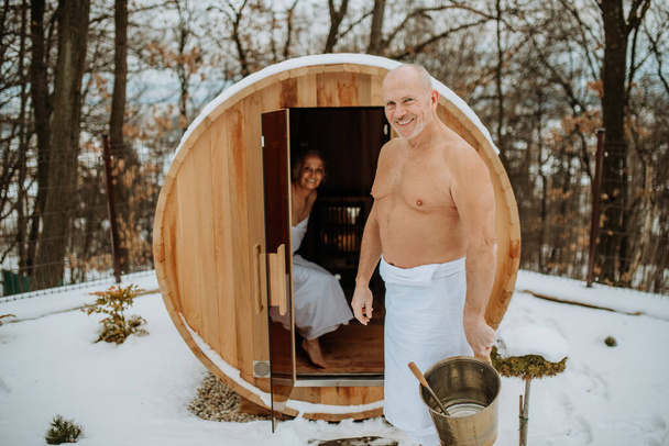 Senior woman in towel with her husband coming out from outdoor sauna during a cold winter day. - Fotó, kép