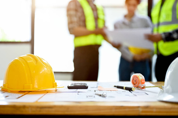 Ingenieur oder Bauarbeiter am Arbeitstisch mit gelbem Hut, Blaupause, Taschenrechner und Maßband auf dem Tisch über verschwommenem Hintergrund von Ingenieuren, die reden - Foto, Bild