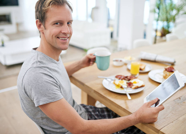 A szombat reggel örömei. Egy jóképű férfi portréja, aki otthon reggelizik a tabletjével. - Fotó, kép