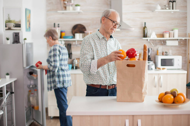 Senior couple arriving from supermarket with grocery bag and unpacking in kitchen. Elderly retired persons enjoying life, spending time helping each other - Photo, Image