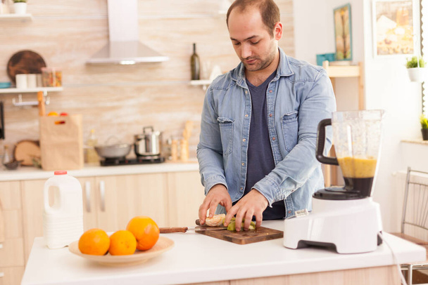 Guy ετοιμάζει θρεπτικό smoothie στην κουζίνα με φρέσκα φρούτα. Υγιής ξέγνοιαστος και χαρούμενος τρόπος ζωής, διατροφή και την προετοιμασία του πρωινού σε ζεστό ηλιόλουστο πρωί - Φωτογραφία, εικόνα