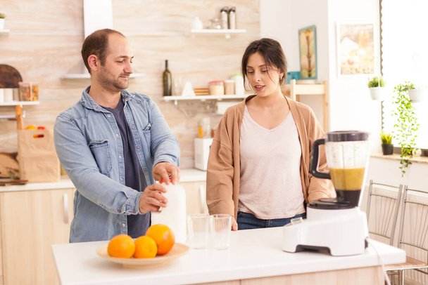 Man opens milk bottle for nutritious smoothie while talking with his girlfriend. Healthy carefree and cheerful lifestyle, eating diet and preparing breakfast in cozy sunny morning - Photo, Image