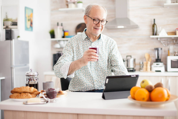 Літній чоловік використовує планшетний ПК на кухні під час сніданку в окулярах. Літня людина з планшетним портативним ПК у пенсійному віці використовує мобільні додатки, сучасні інтернет-інформаційні технології
 - Фото, зображення