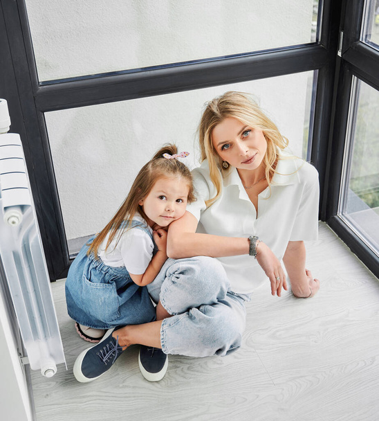 Πορτρέτο της γυναίκας και του μικρού κοριτσιού που κάθεται στο πάτωμα κοντά πανοραμική παράθυρο στο νέο οικογενειακό σπίτι. Μητέρα και παιδί απολαμβάνουν θέα από το παράθυρο στο νεόκτιστο διαμέρισμα. - Φωτογραφία, εικόνα