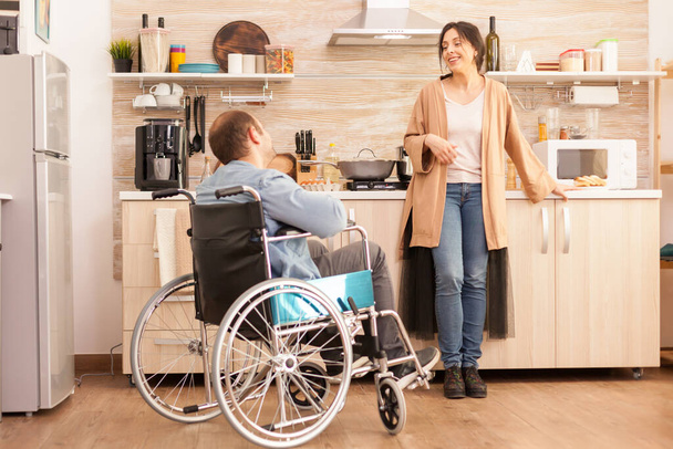 Disabled man sitting in chair and talking with wife while preparing meal. Disabled paralyzed handicapped man with walking disability integrating after an accident. - Fotografie, Obrázek