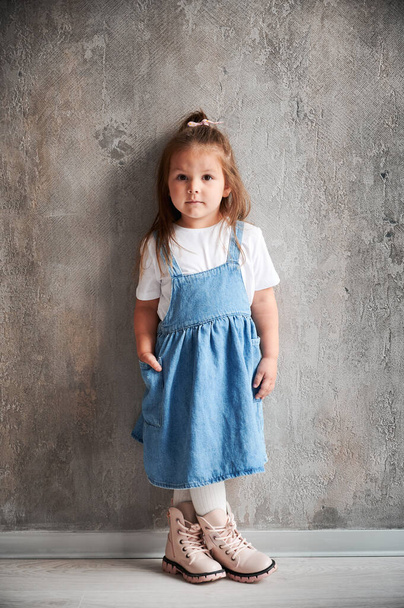 Full length of adorable little girl in denim dress standing against the wall in new home. Cute female child keeping hand in pocket and looking at camera. - Photo, Image
