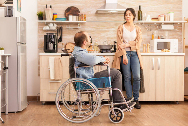 Gehandicapte man in een rolstoel die een gesprek heeft met zijn vrouw in de keuken tijdens het bereiden van voedsel. Gehandicapte, verlamde, gehandicapte, gehandicapte man met een loophandicap die na een ongeval integreert. - Foto, afbeelding