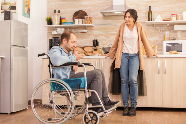Hombre discapacitado en silla de ruedas mirando ensalada hecha por esposa en la cocina. Discapacitado paralítico discapacitado con discapacidad para caminar integrándose después de un accidente. - Foto, imagen