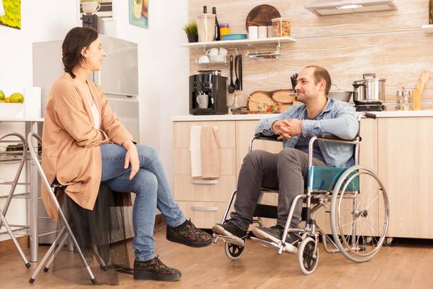 Niepełnosprawny mężczyzna na wózku inwalidzkim patrzący na opiekuńczą żonę w kuchni. Niepełnosprawny sparaliżowany mężczyzna z niepełnosprawnością chodzącą integrujący się po wypadku. - Zdjęcie, obraz