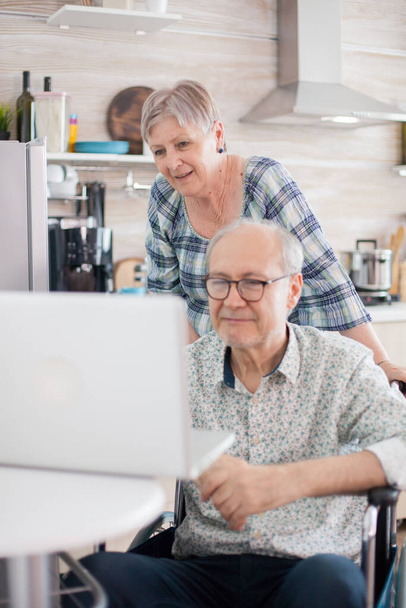 Χαρούμενη ηλικιωμένη γυναίκα σε τηλεδιάσκεψη με την οικογένειά της. Ο ανάπηρος ηλικιωμένος σε αναπηρική καρέκλα και η γυναίκα του έχουν τηλεδιάσκεψη στο λάπτοπ στην κουζίνα. Παράλυτος γέρος και η σύζυγός του έχουν ένα online - Φωτογραφία, εικόνα