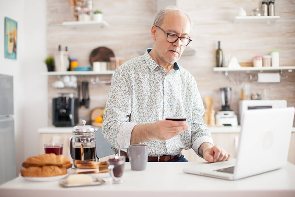 Starszy mężczyzna robi zakupy online rano ze swojej kuchni. Emeryt płaci online za pomocą karty kredytowej i aplikacji z laptopa. Emeryt i rencista korzystający z płatności internetowej - Zdjęcie, obraz