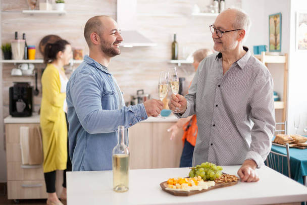 Szczęśliwy ojciec i syn brzęczenie kieliszki wina w kuchni podczas lunchu z rodziną. - Zdjęcie, obraz