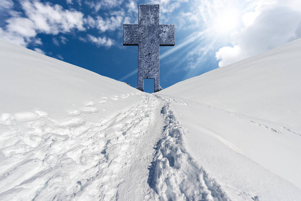 Grande croce religiosa in pietra con una porta e un sentiero con impronte sulla neve in un paesaggio montano contro un cielo blu con nuvole, raggi di sole e spazio copia. - Foto, immagini