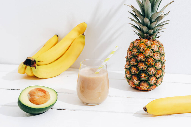 Still life with fruits, banana, pineapple and avocado smoothie on white wooden table with long shadows. Minimal detox diet concept, summer vitamin drink. - Photo, Image