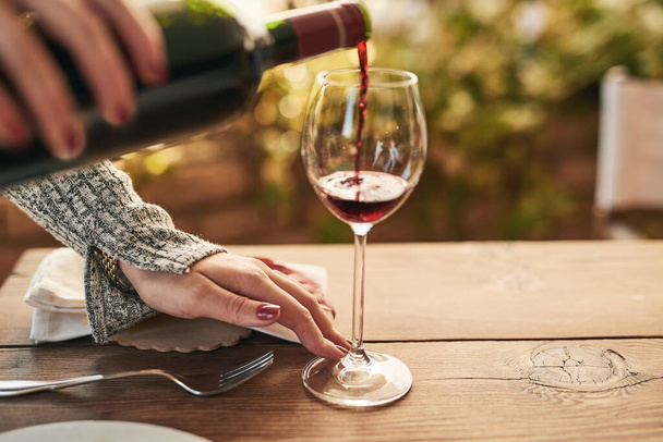 Glas, fles en hand van de vrouw gieten wijn aan een tafel, rustig en tevreden onder het genot van vrije tijd op een patio. Rode wijn, handen en dame ontspannen met luxe drankje, genieten van een ontspannen weekend alleen thuis. - Foto, afbeelding