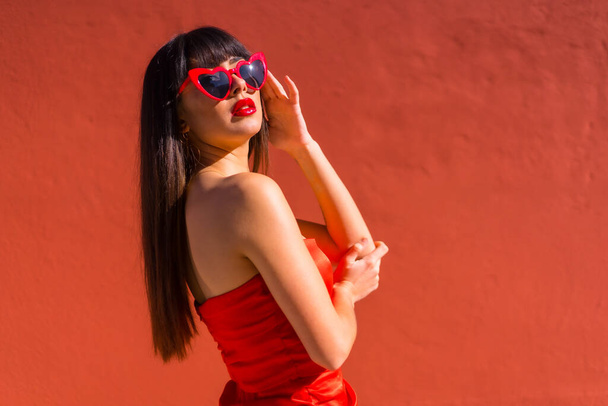 Chica morena sobre un fondo rojo con gafas de corazones y vestido ajustado rojo, Día de San Valentín - Foto, Imagen