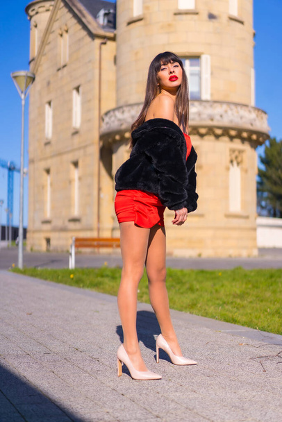 Mladá žena v červených šatech v krásném zámku, módně pózuje a usmívá se v černé bundě - Fotografie, Obrázek
