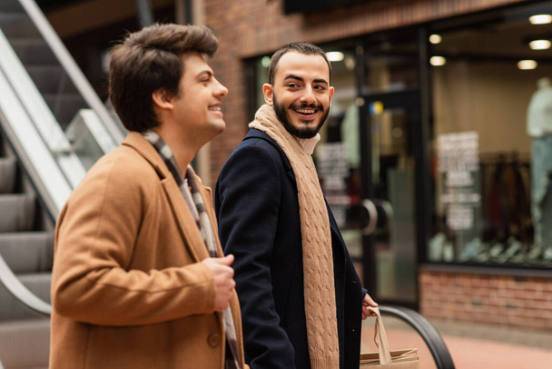 Fröhlicher bärtiger Mann mit Einkaufstüten schaut schwulen Partner auf der Straße an - Foto, Bild