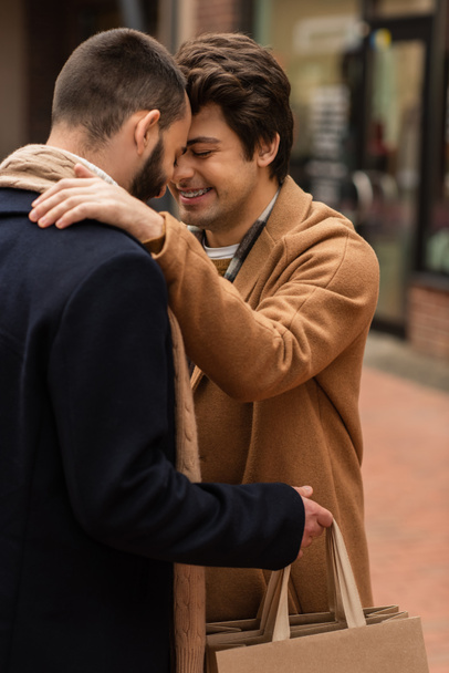 hombre gay de moda con los ojos cerrados sonriendo cerca de novio sosteniendo bolsas de compras en la calle urbana - Foto, imagen