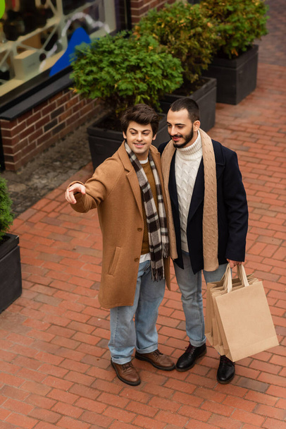 Blick aus der Vogelperspektive auf einen lächelnden schwulen Mann, der mit dem Finger zeigt und in der Nähe seines Freundes mit Einkaufstüten wegsieht - Foto, Bild
