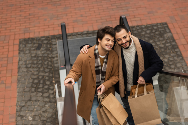 high angle view of bearded gay man embracing trendy boyfriend while holding shopping bags on escalator - 写真・画像