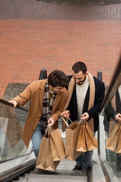 θέα υψηλής γωνίας των κομψών και χαρούμενων ομοφυλόφιλων ανδρών που αναζητούν τσάντες για ψώνια στις κυλιόμενες σκάλες - Φωτογραφία, εικόνα