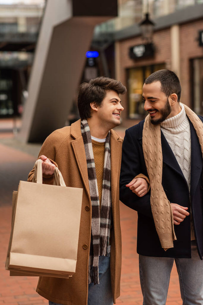 joyful gay man holding shopping bags and smiling at bearded boyfriend on blurred street - Fotó, kép