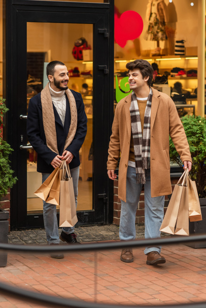 full length of cheerful gay men in stylish clothes holding shopping bags near store on city street - Foto, afbeelding