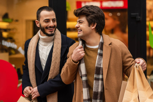 smiling gay man holding shopping bags and adjusting scarf near trendy bearded boyfriend and blurred showcase - Photo, Image