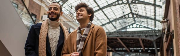 young and fashionable gay men smiling and looking away under transparent roof outdoors, banner - Photo, Image