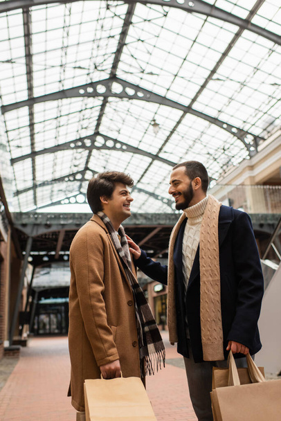 cheerful gay men in trendy outfits holding shopping bags and looking at each other under transparent roof on street - Photo, Image
