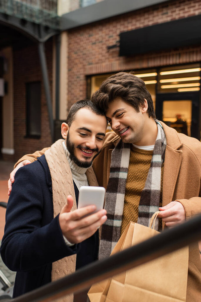 gelukkig gay man met winkelen tassen omarmen trendy bebaarde vriend terwijl op zoek naar mobiele telefoon - Foto, afbeelding