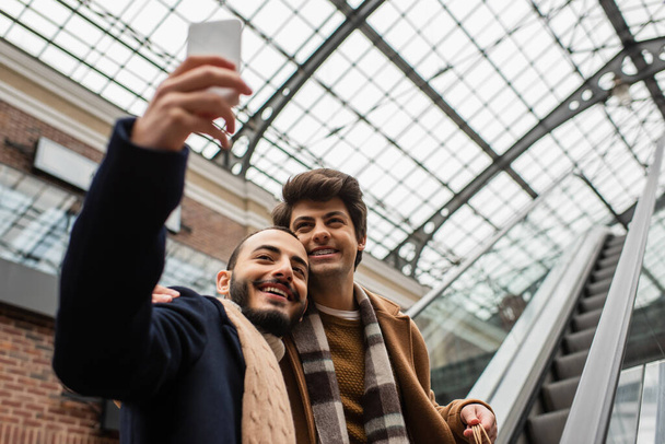 bärtiger Schwuler macht Selfie mit jungem Freund in kariertem Schal auf Rolltreppe - Foto, Bild