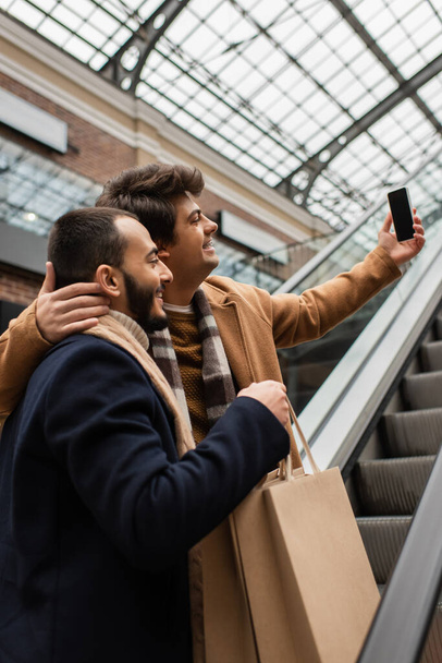 junger Mann umarmt Hals eines bärtigen und trendigen Freundes mit Einkaufstaschen und macht Selfie auf Rolltreppe - Foto, Bild