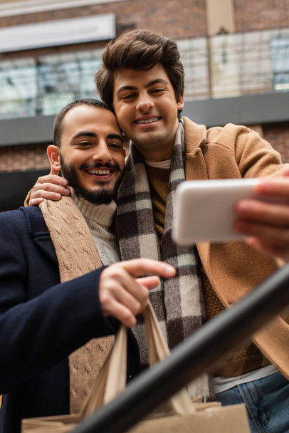 smiling gay man embracing bearded boyfriend with shopping bags while taking selfie on blurred mobile phone - Фото, зображення