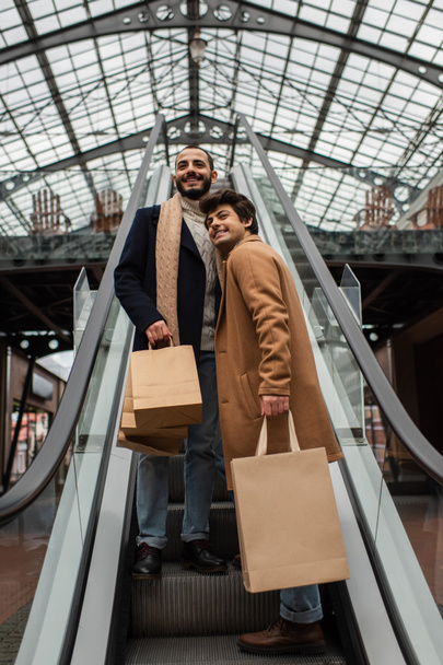 low angle view of cheerful gay men in stylish casual clothes holding shopping bags and looking away on escalator - Photo, Image