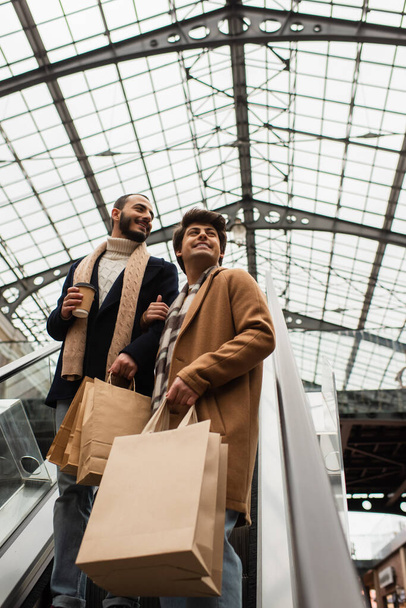 low angle view of trendy and happy gay couple with shopping bags and paper cup looking away on escalator - Photo, Image