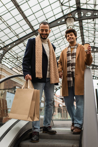 mosolygós meleg férfiak divatos ruhák áll bevásárló táskák és papír csésze mozgólépcsőn - Fotó, kép