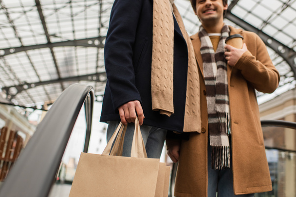 Bulanık erkek arkadaşının yanında alışveriş torbalarıyla gezen ve yürüyen merdivende içki içen modaya uygun bir gay. - Fotoğraf, Görsel