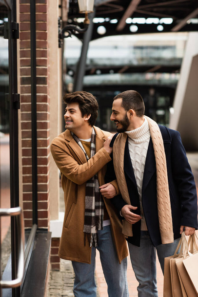 cheerful and trendy gay men with shopping bags looking at showcase on city street - Foto, imagen