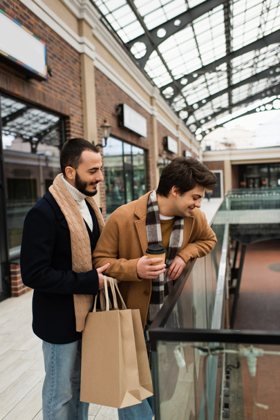 gay man with paper cup looking down near glass fence and bearded boyfriend with shopping bags - Foto, Imagen