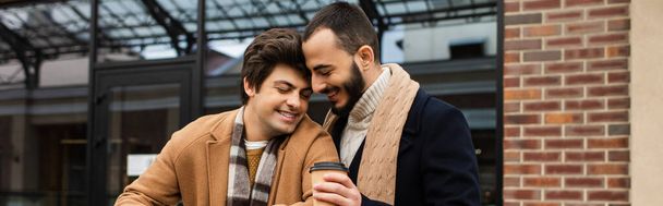 喜ばしい髭を生やした男がコーヒーを持って街のゲイのパートナーの近くに行き - 写真・画像
