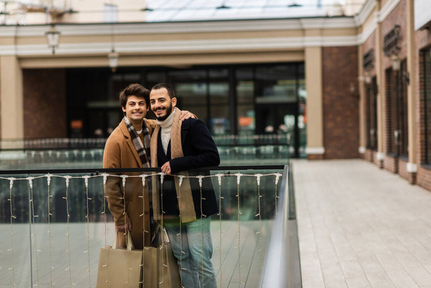 trendy i szczęśliwy gej mężczyźni z zakupy torby stojąc w pobliżu szkło ogrodzenie i zamazany budynek - Zdjęcie, obraz