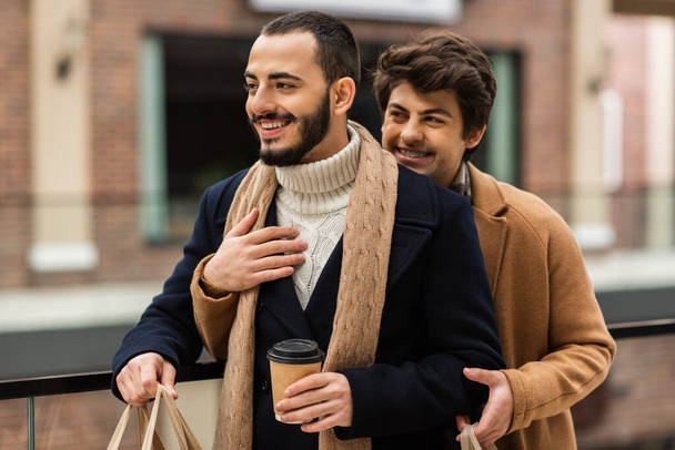bearded gay in coat and scarf standing with takeaway drink near boyfriend embracing him outdoors - Photo, Image