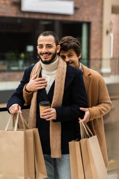 bearded gay man in black coat holding shopping bags and paper cup near young boyfriend hugging him outdoors - Фото, изображение