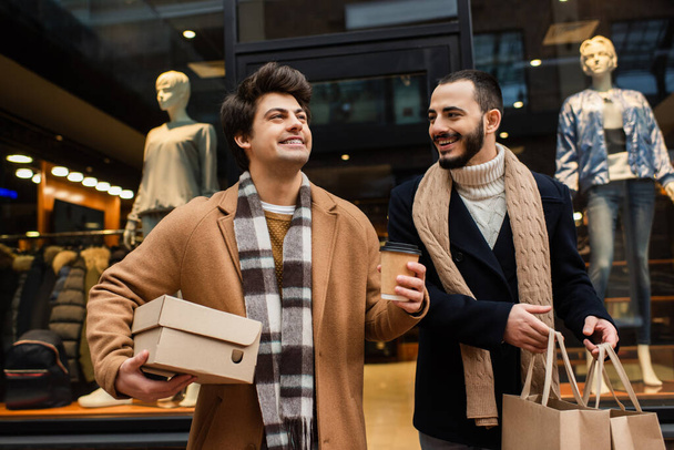 Alışveriş torbalarıyla sakallı eşcinsel adam elinde kağıt bardakla mutlu erkek arkadaşına bakıyor ve vitrinin yanındaki mankenli ayakkabı kutusuna bakıyor. - Fotoğraf, Görsel
