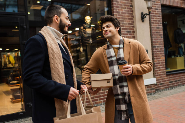 felice gay uomo con shoebox e take away bere guardando barbuto fidanzato con shopping bags vicino offuscata negozio - Foto, immagini