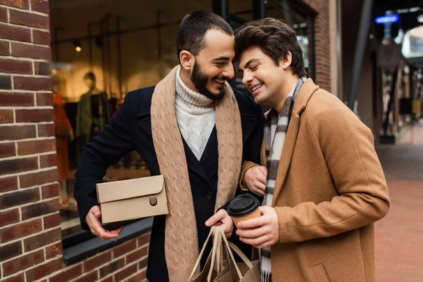 happy gay couple with purchases and paper cup standing near blurred showcase on city street - Photo, Image