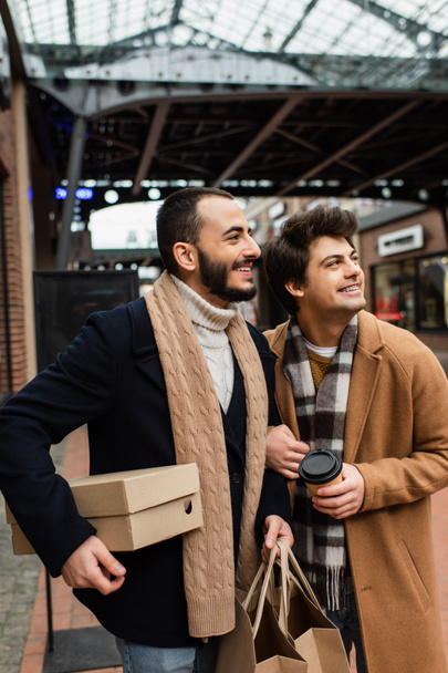 trendy gay paar met aankopen en koffie te gaan glimlachen en weg te kijken op wazig straat - Foto, afbeelding