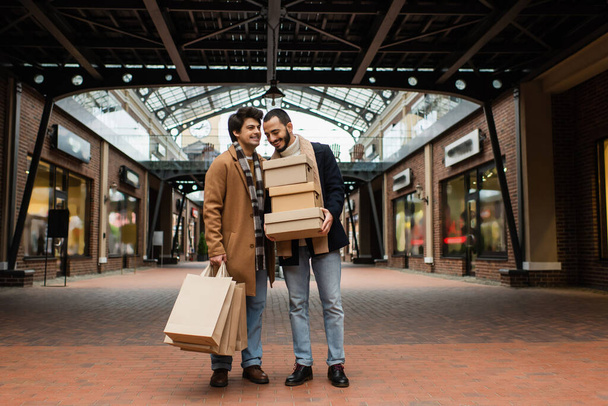 都会の通りに店があるビルの近くにショッピングバッグや靴箱を持つ流行のゲイカップルの完全な長さ - 写真・画像